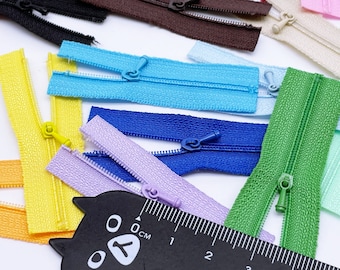 Può includere: Assortimento di chiusure a zip colorate in vari colori, tra cui rosa, blu, verde, giallo e viola. Le cerniere sono disposte su una superficie bianca con un righello nero contrassegnato in centimetri.