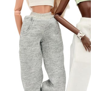 May include: Two dolls in casual attire. One doll wears a white ribbed crop top and grey sweatpants. The other doll is dressed in a light green crop top and white trousers. The dolls are set against a white backdrop.
