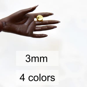 以下が含まれることがあります： 3mmの丸い金色のスタッドピアスのクローズアップ。ピアスはマネキンの手によって保持されています。手の下に「3mm」と「4色」のテキストが表示されています。