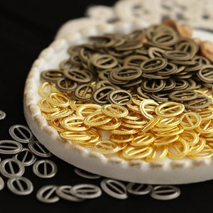 May include: A white ceramic dish filled with gold, bronze, and silver metal rings. The dish is on a white lace doily. The rings are scattered on a black surface.