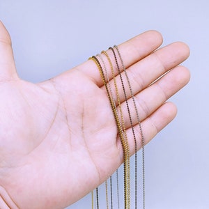 May include: A close-up of a hand holding a variety of silver, gold, and brown bead chains. The chains are all the same length and are arranged in a neat row.