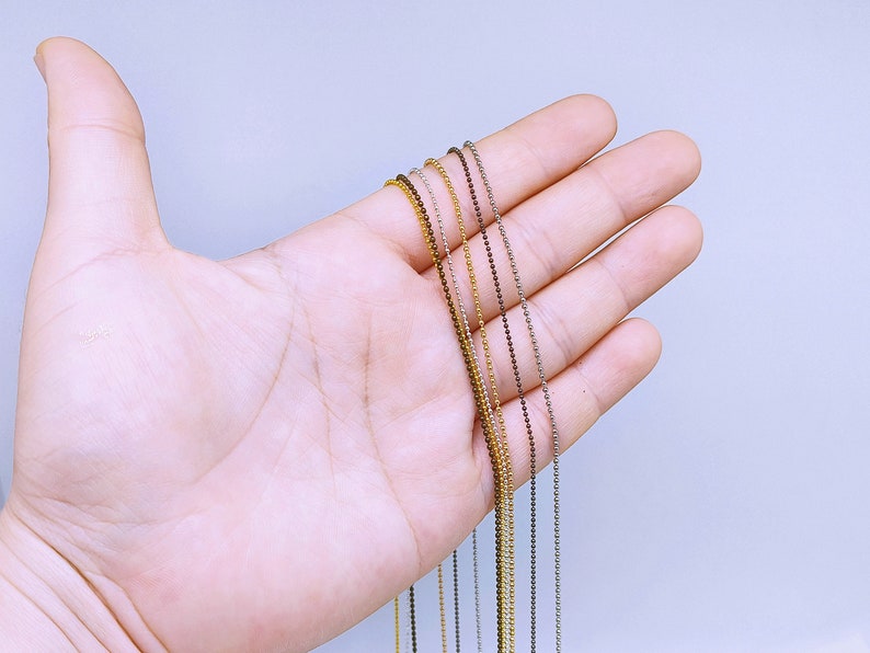May include: A close-up of a hand holding a variety of silver, gold, and brown bead chains. The chains are all the same length and are arranged in a neat row.