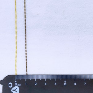 May include: Two chains of beads, one gold and one brown, are laid out on a white surface next to a black ruler marked in centimeters. The gold chain is on the left and the brown chain is on the right. The chains are both about 6 centimeters long.