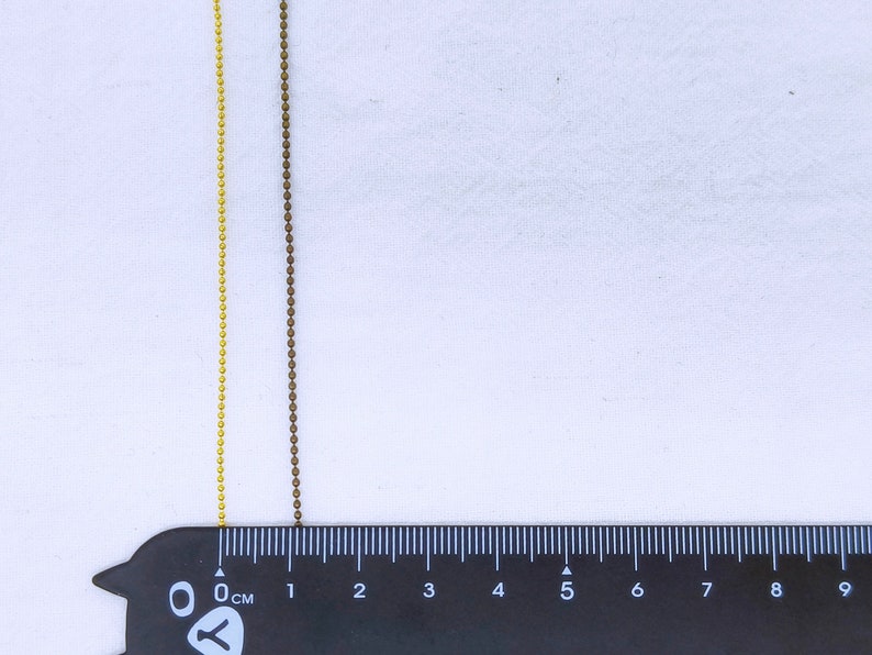 May include: Two chains of beads, one gold and one brown, are laid out on a white surface next to a black ruler marked in centimeters. The gold chain is on the left and the brown chain is on the right. The chains are both about 6 centimeters long.
