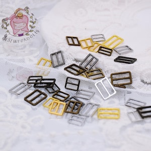 May include: Assortment of metal slider buckles in gold, silver, and bronze finishes. The buckles are rectangular with a center opening and are scattered on a white lace background.