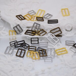 May include: A collection of silver, gold, and bronze metal slider buckles. The buckles are all the same size and shape, but they are different colors. They are arranged in a random pattern on a white surface.