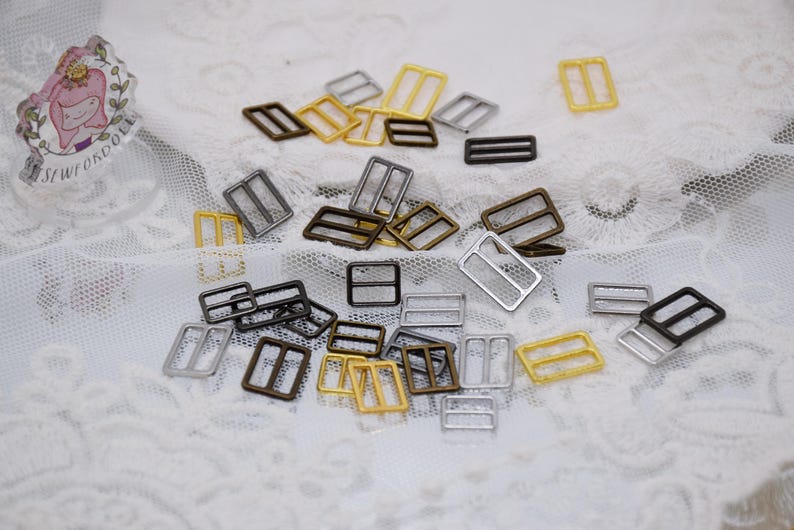 May include: A collection of silver, gold, and bronze metal slider buckles. The buckles are all the same size and shape, but they are different colors. They are arranged in a random pattern on a white surface.