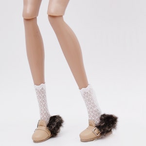 Pode incluir: Um par de sapatos de boneca cor de areia com acabamento de pele e fivelas. Os sapatos são usados por uma boneca com meias de rede brancas.