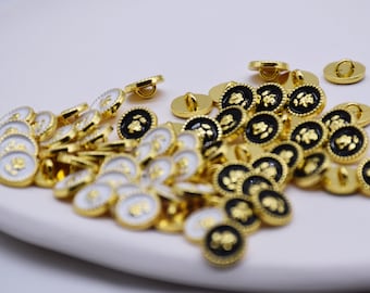 Può includere: Un grande mucchio di bottoni dorati con un designo in rilievo. Alcuni bottoni sono neri con accenti dorati, mentre altri sono bianchi con accenti dorati.