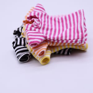 Op de afbeelding: Een stapel van drie gestreepte poppenhemden in roze en wit, geel en oranje en zwart en wit.