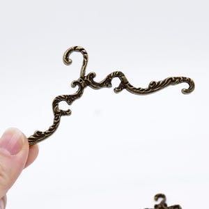 Puede incluir: Herraje decorativo de metal de color bronce antiguo y ornamentado. La pieza decorativa tiene un diseño curvo y asimétrico con volutas y adornos. El herraje se sostiene en una mano sobre un fondo blanco. Probablemente para muebles o proyectos de artesanía.