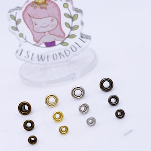 May include: Assortment of metal eyelets in various sizes and finishes, including gold, silver, and bronze. The eyelets are arranged on a white surface.