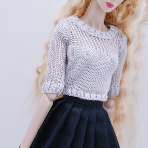 May include: A doll wearing a white crocheted sweater and a black pleated skirt. The doll has long blonde hair and blue eyes.
