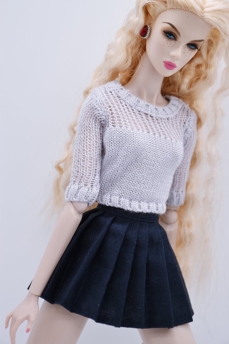 May include: A doll wearing a white crocheted sweater and a black pleated skirt. The doll has long blonde hair and blue eyes.
