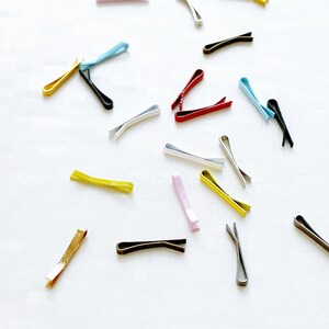 May include: Assortment of colourful metal hair clips in various shapes and sizes. The clips are arranged on a white surface.
