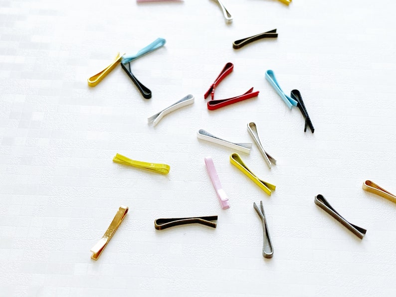 May include: Assortment of colourful metal hair clips in various shapes and sizes. The clips are arranged on a white surface.