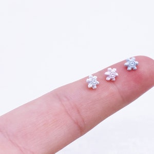 May include: Three small snowflake-shaped charms in different colours: light blue, pink, and white. The charms are on a finger.