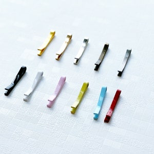 May include: Assortment of 12 hair clips in various colours, including black, gold, silver, white, pink, yellow, blue, and red. The clips are all the same size and shape, and they are arranged in a row on a white surface.