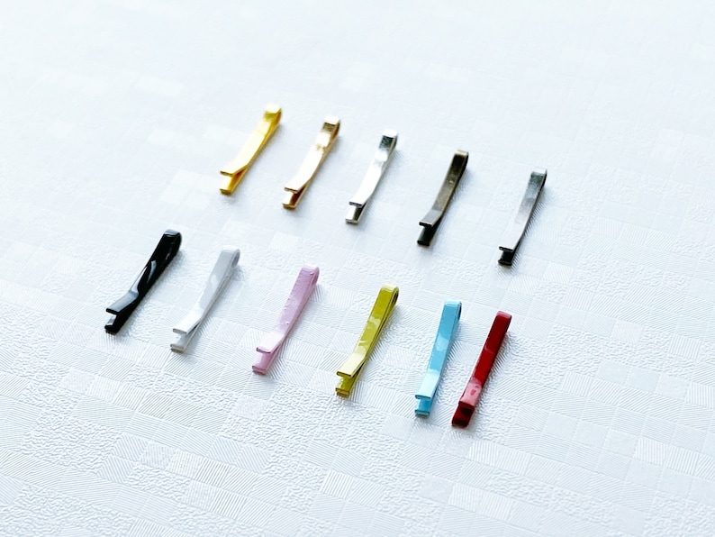 May include: Assortment of 12 hair clips in various colours, including black, gold, silver, white, pink, yellow, blue, and red. The clips are all the same size and shape, and they are arranged in a row on a white surface.