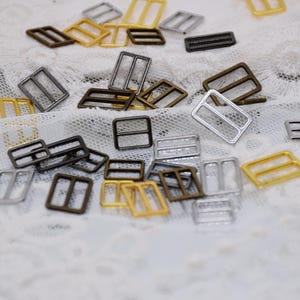May include: Assortment of metal slider buckles in gold, silver, and bronze finishes. The buckles are rectangular with two parallel bars across the center.
