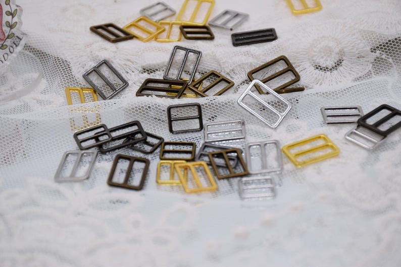 May include: Assortment of metal slider buckles in gold, silver, and bronze finishes. The buckles are rectangular with two parallel bars across the center.