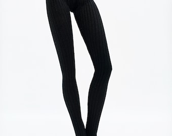 Può includere: Leggings neri a coste. I leggings hanno una vita alta e una vestibilità slim, con una trama a coste verticali. Il tessuto sembra essere di peso medio, adatto per le temperature più fresche. I leggings sono mostrati su una bambola.