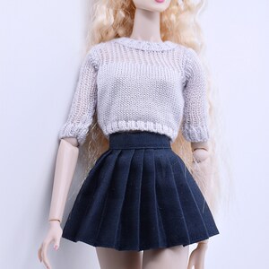 May include: A doll wearing a white knitted sweater and a black pleated skirt.