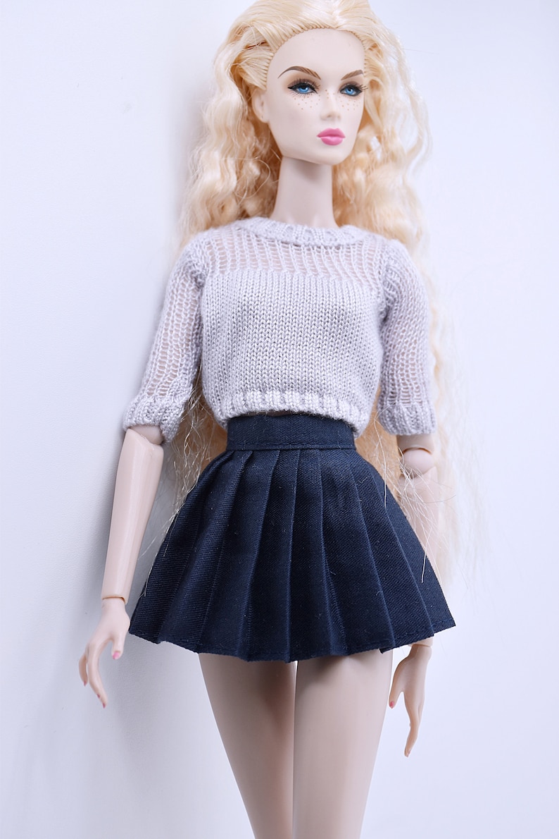 May include: A doll wearing a white knitted sweater and a black pleated skirt.