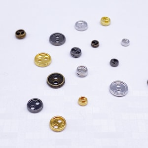 May include: Assortment of two-hole buttons in various colors, including gold, silver, and black. The buttons are arranged on a white surface.