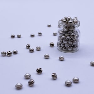 May include: A small glass jar filled with silver-colored, pearl-topped beads. Some beads are scattered around the jar on a white surface. The beads have a metallic finish and a small, decorative design on the back.