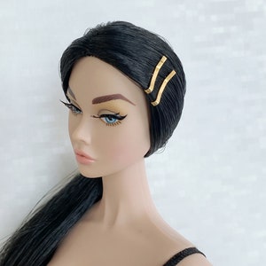 May include: A black haired doll with blue eyes wearing a black top and gold hair clips.
