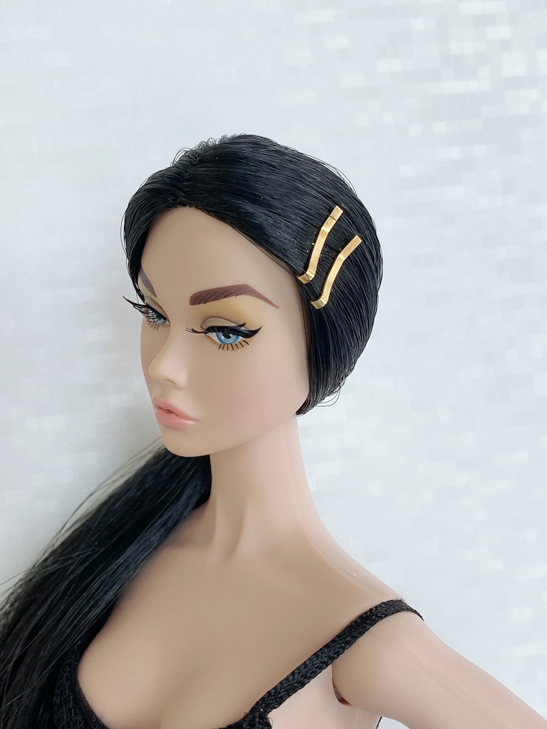 May include: A black haired doll with blue eyes wearing a black top and gold hair clips.