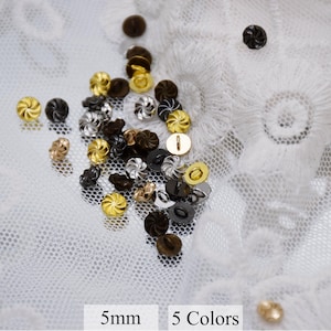 May include: Assortment of 5mm metal buttons in five colors: gold, silver, black, brown, and copper. The buttons have a swirl design and are displayed on a white lace background.