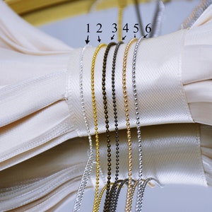 May include: Six different types of chain, numbered 1 through 6, are displayed on a white fabric background. The chains are made of metal and are in various colors, including gold, silver, and black.