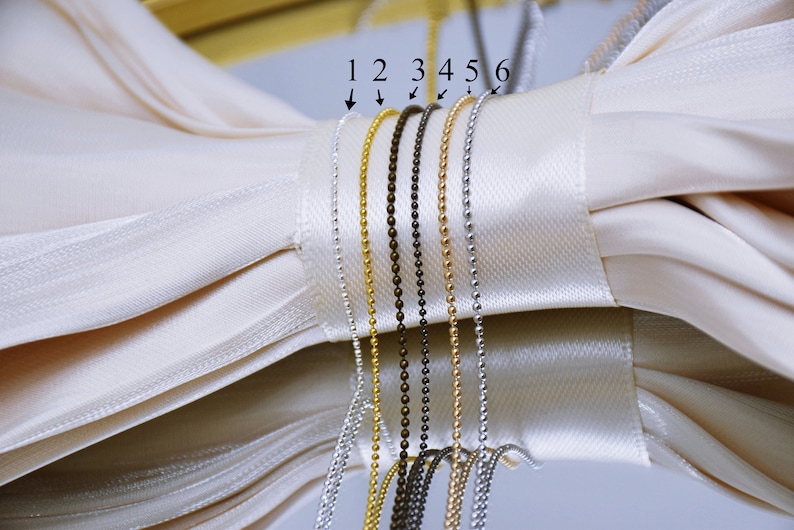 May include: Six different types of chain, numbered 1 through 6, are displayed on a white fabric background. The chains are made of metal and are in various colors, including gold, silver, and black.