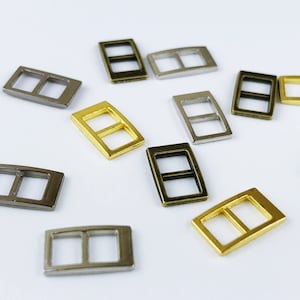 May include: Assortment of metal slider buckles in gold, silver, and black finishes. The buckles have two holes and are rectangular in shape.