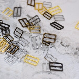 May include: Assortment of metal slider buckles in silver, gold, and bronze finishes. The buckles are rectangular with two holes on each side. They are scattered on a white lace background.