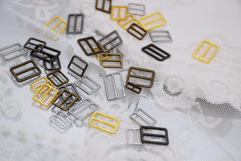 May include: Assortment of metal slider buckles in silver, gold, and bronze finishes. The buckles are rectangular with two holes on each side. They are scattered on a white lace background.
