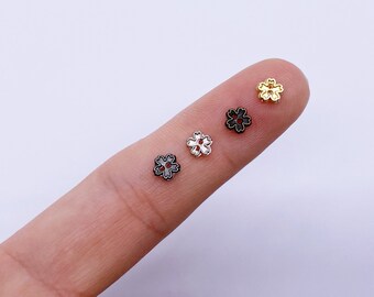 May include: Four small flower-shaped metal beads in different colors: gold, silver, black, and white. The beads are shown on a finger.