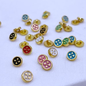 以下が含まれることがあります： さまざまな色のエナメルデザインが施された、小さく丸い金色のボタンの詰め合わせ。ボタンの中央には、4つに分かれた星のデザインがあります。