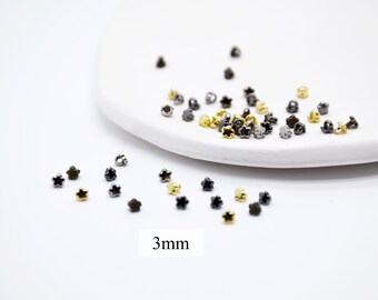 May include: A collection of small, star-shaped beads in gold and dark gray hues. The beads are scattered on a white surface, with some resting on a white, oval-shaped dish. The text "3mm" is visible.