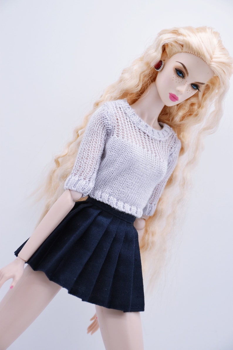 May include: A doll wearing a white knitted sweater and a black pleated skirt. The doll has long blonde hair and blue eyes.