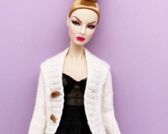 May include: A fashion doll with blonde hair styled in a high bun, wearing a white knit cardigan with wooden toggle closures over a black dress. The doll has red lipstick and is set against a purple background.