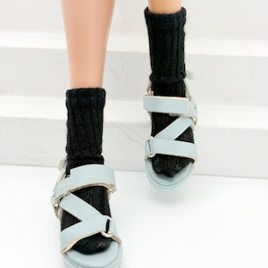 May include: Pale blue sandals with silver buckles and black socks. The sandals feature a criss-cross strap design and a thick sole. The socks are ribbed and reach mid-calf. The sandals are on a white background.