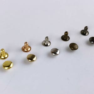 May include: Assortment of metal rivets in various finishes, including gold, silver, bronze, and copper. The rivets are small and round, with a flat head and a pointed shank.