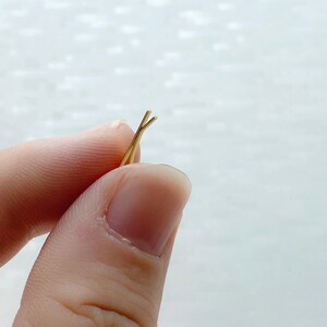 May include: A pair of gold bobby pins, also known as hair clips, are held in a person's hand. The bobby pins are crossed at the top.