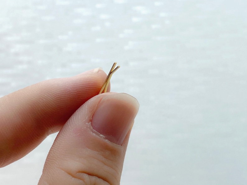 May include: A pair of gold bobby pins, also known as hair clips, are held in a person's hand. The bobby pins are crossed at the top.