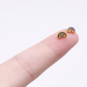 May include: Two small rainbow-shaped earrings with gold-colored borders. One earring features a black, yellow, and blue rainbow design, while the other has a red, orange, and blue rainbow design. The earrings are small, likely for pierced ears.