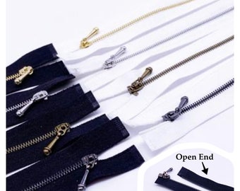May include: Assortment of white and black zipper tapes with different colored metal zipper pulls. The zipper tapes are laid out in a row, with the black tapes on top of the white tapes. The zipper pulls are in various colors, including gold, silver, and bronze. The image also shows a close-up of an open-ended black zipper tape.