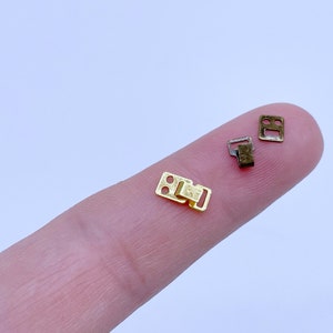 May include: Three small metal clasps, two gold and one silver, are shown on a finger. The clasps are rectangular with two small holes in each.
